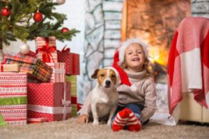 A dog sits with a young girl under a Christmas Tree